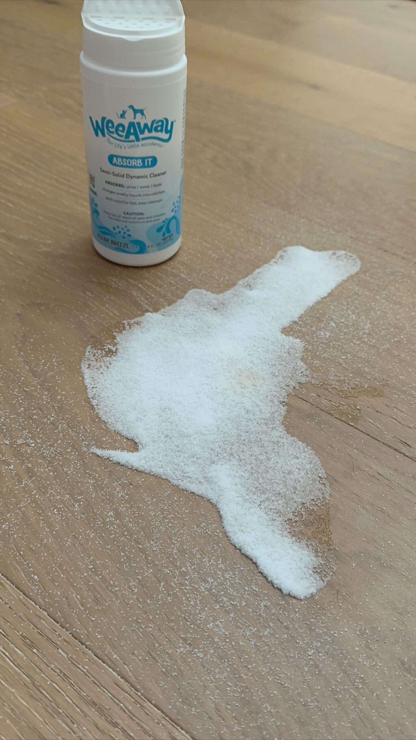 Wee Away Absorb It canister behind a liquid puddle. The puddle was sprinkled with Absorb It and the powder is congealing the liquid so it is easier to clean up.