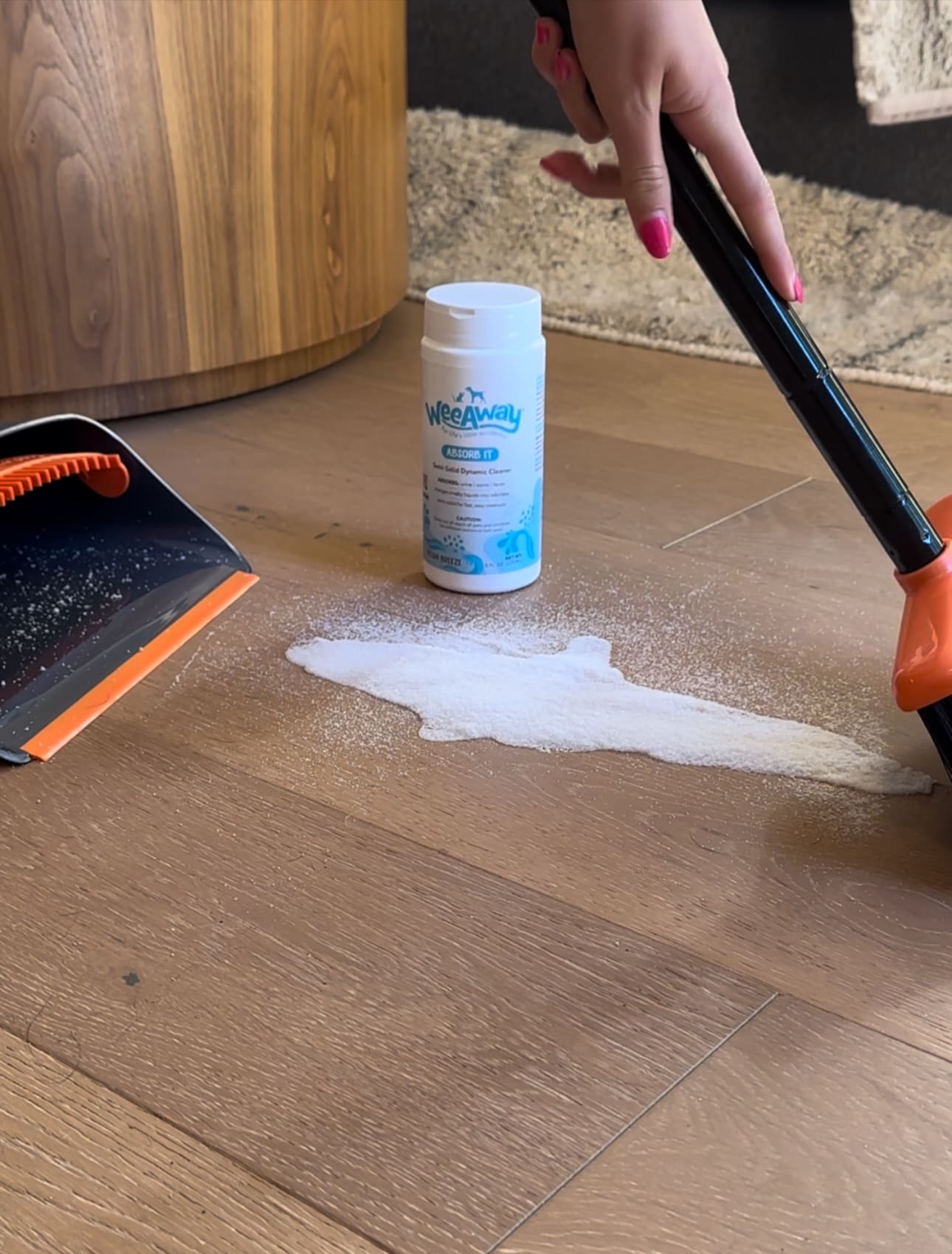 Wee Away Absorb It canister behind a liquid puddle. The puddle was sprinkled with Absorb It and the powder is congealed and being swept into a dustpan.
